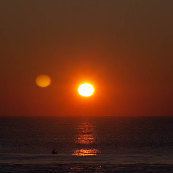 Coucher de soleil sur la mer. Ambiance très rouge. On devinne un surfeur à l'eau attendant une vague.